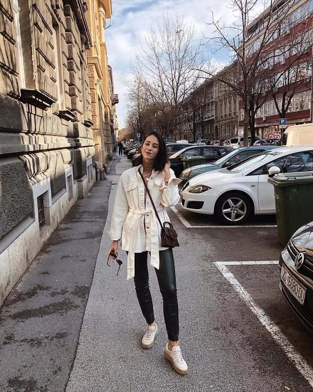 Woman walking on a city street wearing a beige turtleneck, utility jacket, black leather pants, sneakers