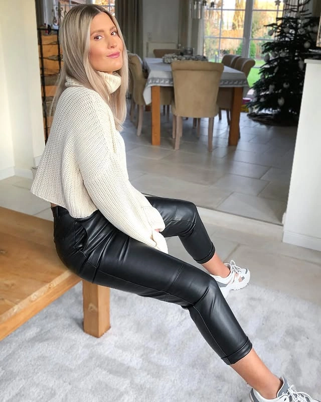 Woman sitting on a wooden bench indoors, wearing a beige cropped turtleneck sweater, black leather leggings