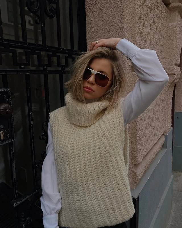  Woman leaning against a metal gate, wearing a beige chunky knit turtleneck vest layered over a white shirt