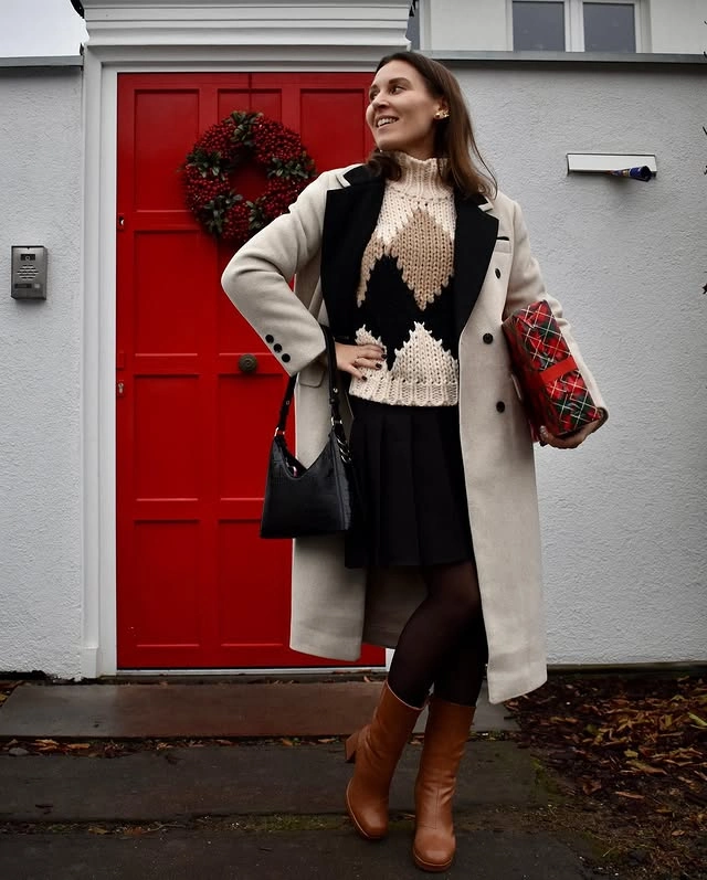 Woman standing in front of a red door adorned with a holiday wreath, wearing a beige argyle turtleneck sweater, black pleated skirt