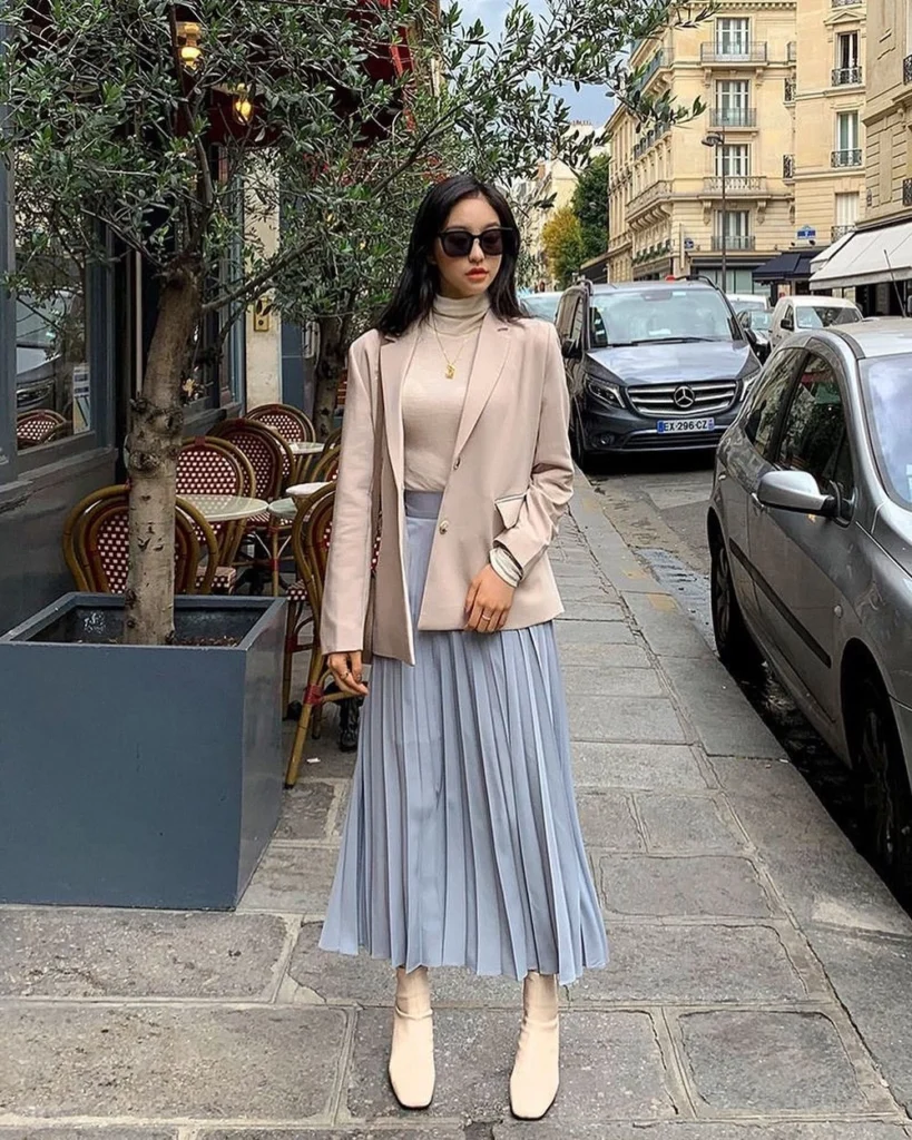 Woman dressed in a beige turtleneck, blazer, pastel blue pleated skirt, and beige ankle boots