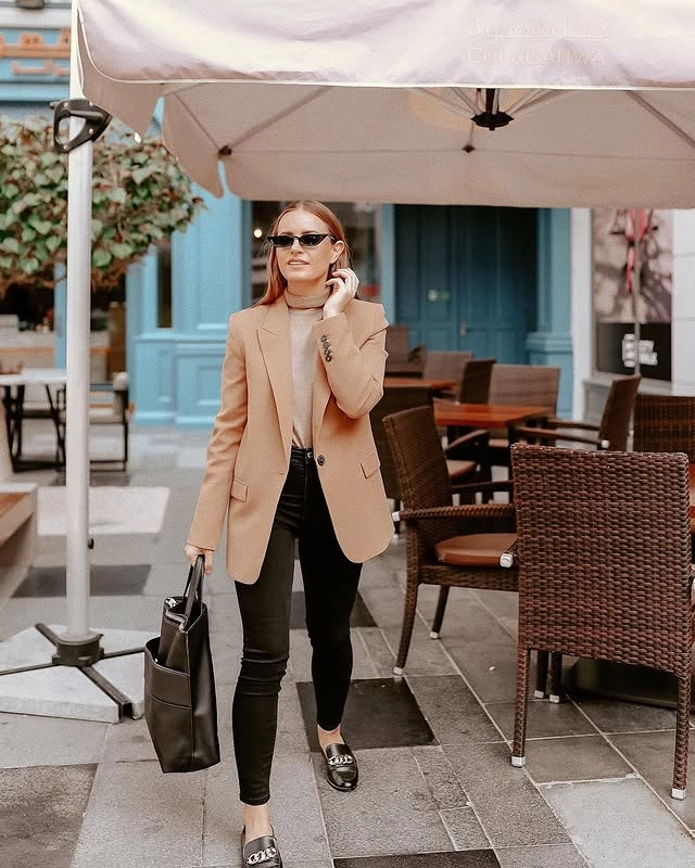 Woman wearing a beige turtleneck, camel blazer, black skinny jeans, and black loafers, accessorized with a black tote bag and sunglasses