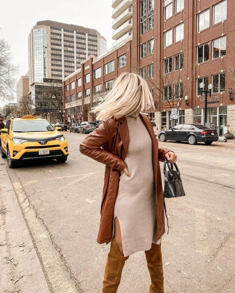 Woman wearing a beige ribbed turtleneck sweater dress with side slits, a brown leather trench coat, knee-high suede boots